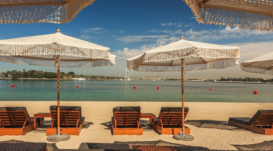 Beach with wooden lounge chairs under fringed umbrellas facing a calm sea, with a distant cityscape and clear blue sky.