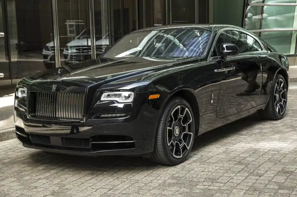 A sleek, black luxury car with a shiny finish is parked on a cobblestone surface in an urban setting.