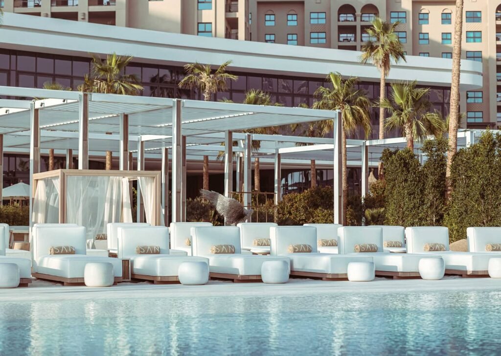 Palm Jumeirah Night Swim: Poolside lounge area with white cushioned seating, palm trees, and a modern building in the background.