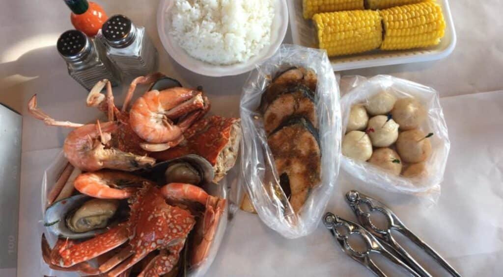 A seafood platter with crabs, shrimp, and mussels, alongside rice, corn on the cob, and fish slices, with sauce and utensils.