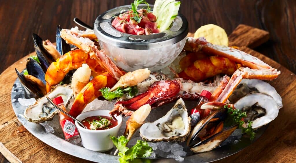 A seafood platter with oysters, crab legs, shrimp, and mussels, garnished with greens and served with sauce and lime wedges.