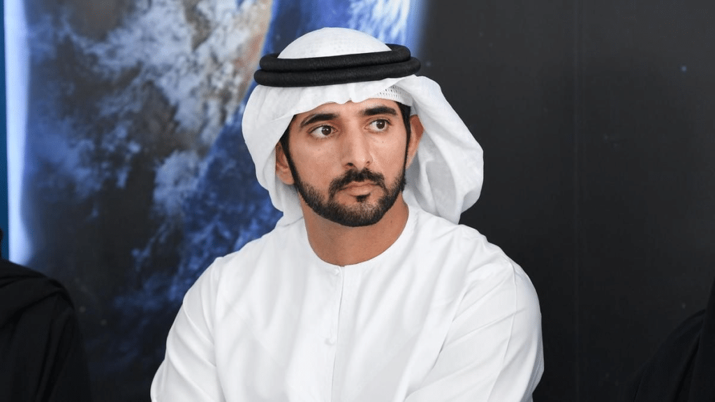 Sheikh Hamdan bin Mohammed bin Rashid Al Maktoum in a white traditional Middle Eastern outfit, including a keffiyeh, with a large image of Earth in the background.