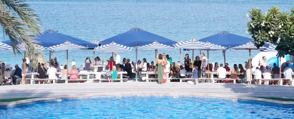 People enjoying a beachside gathering under blue and white striped umbrellas near a clear blue swimming pool.