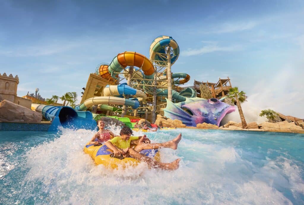 Children ride down a colorful water slide on an inflatable raft, splashing into the pool below against a backdrop of twisting slides.