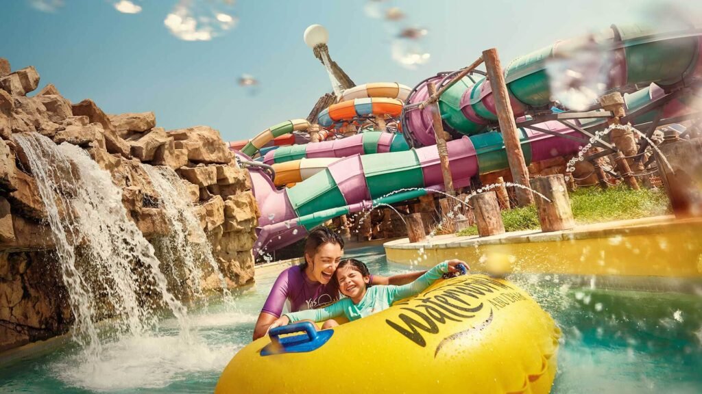 A person and a child are joyfully riding a yellow inflatable raft on a water slide ride with colorful tubes and splashes around.