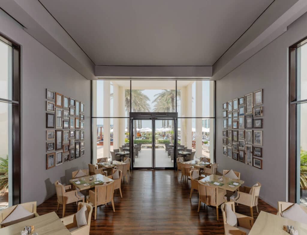 A modern restaurant with wooden floors, tables, and chairs, featuring large windows and picture frames on the walls, overlooking an outdoor seating area and palm trees.