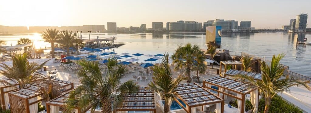 Sunset over a waterfront beach club with palm trees, blue umbrellas, lounge chairs, and city buildings across the water