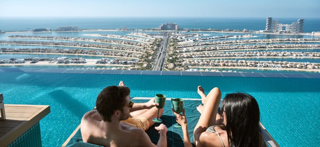 A couple relaxes on lounge chairs by an infinity pool, overlooking a coastal cityscape with palm-shaped islands.