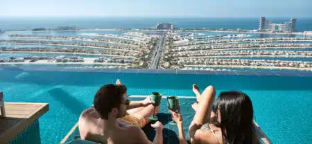 A couple relaxes on lounge chairs by an infinity pool, overlooking a coastal cityscape with palm-shaped islands.