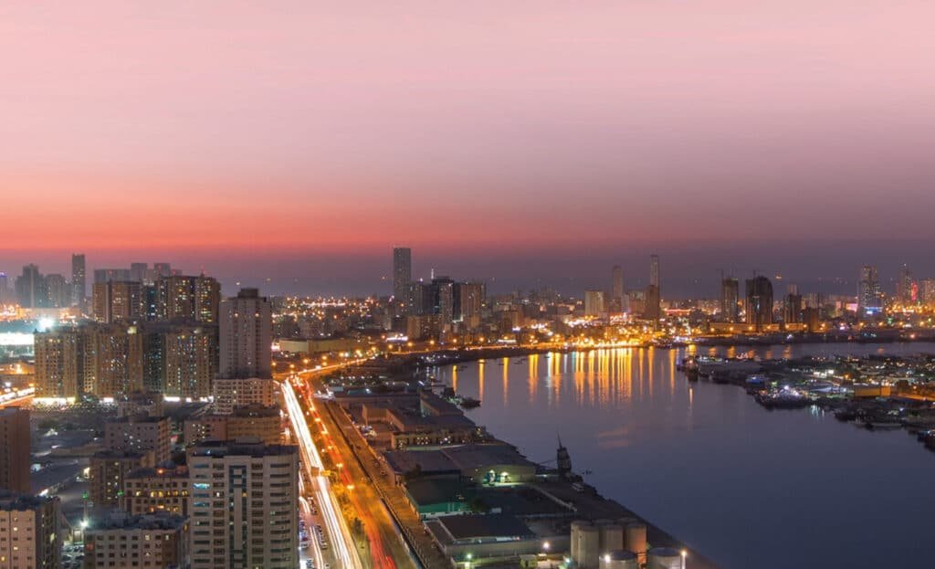 City skyline at dusk with glowing lights reflected on a river under a pink sunset sky