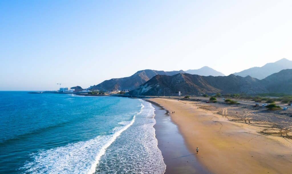 Aerial view of a curved sandy beach with turquoise waves, rocky mountains, and a few people walking along the shore