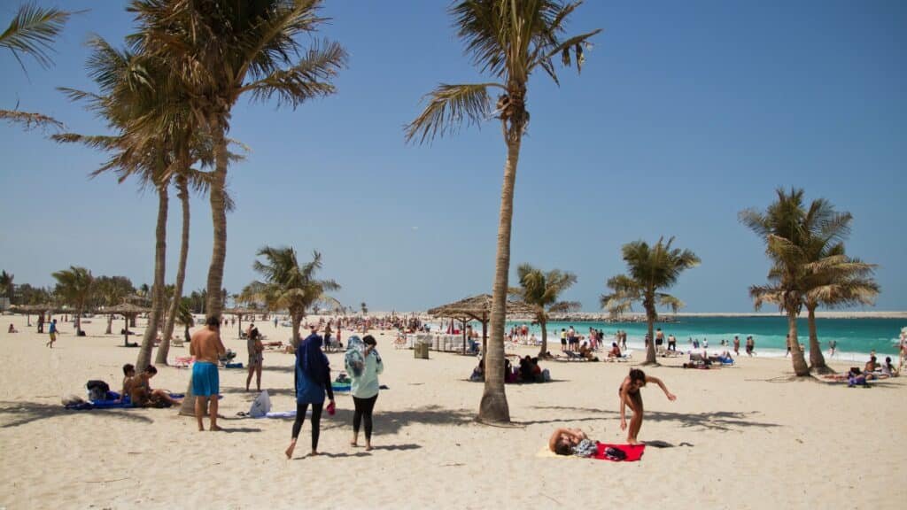 A sunny Al Mamzar Beach with palm trees and people relaxing, sunbathing, and swimming in the ocean.