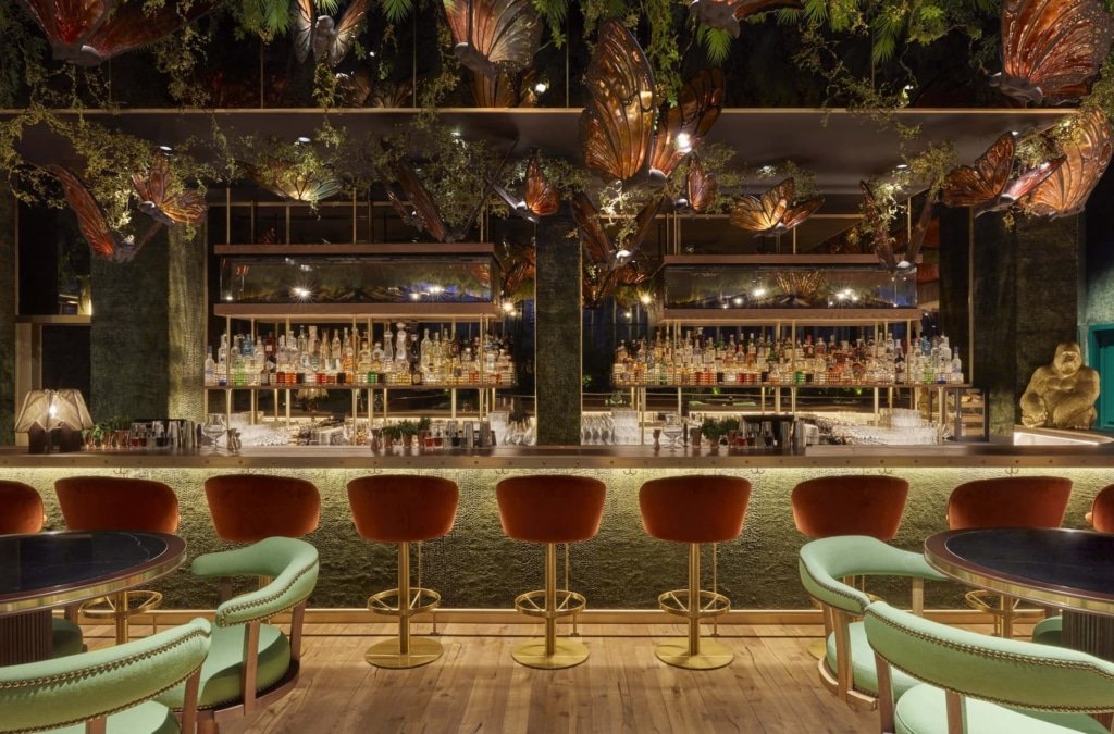 Luxurious bar interior with a row of red velvet stools, a well-stocked backlit bar, and decorative plants hanging overhead.
