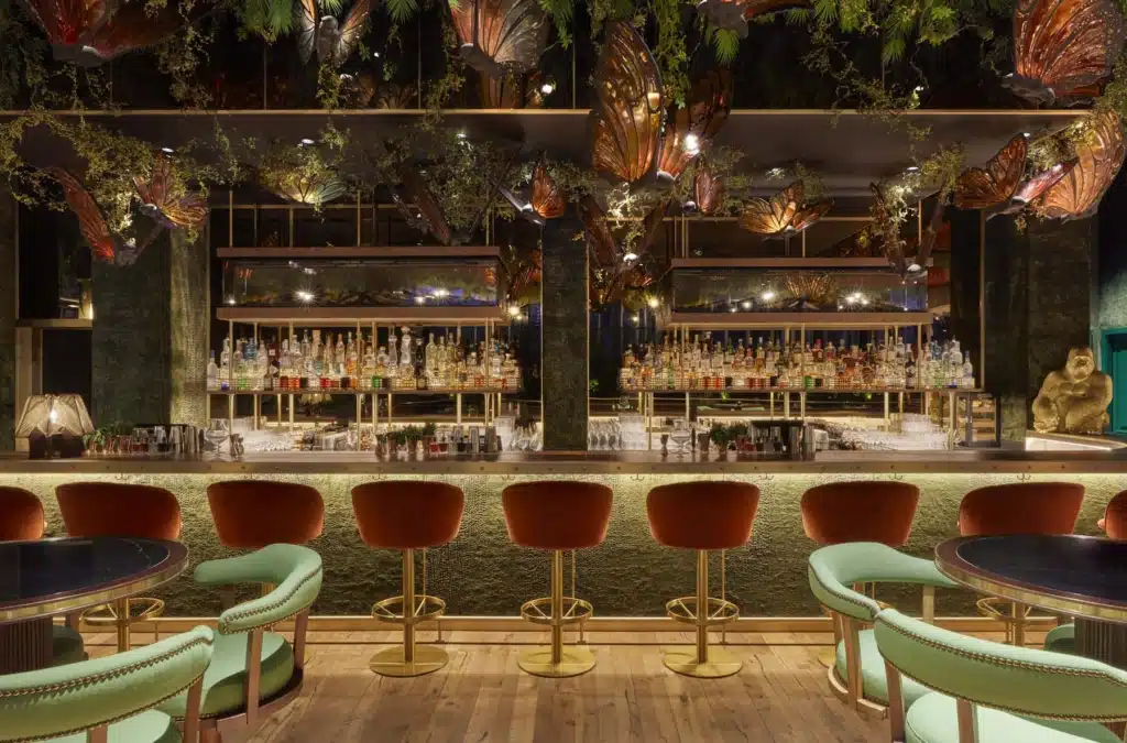 Luxurious bar interior with a row of red velvet stools, a well-stocked backlit bar, and decorative plants hanging overhead.