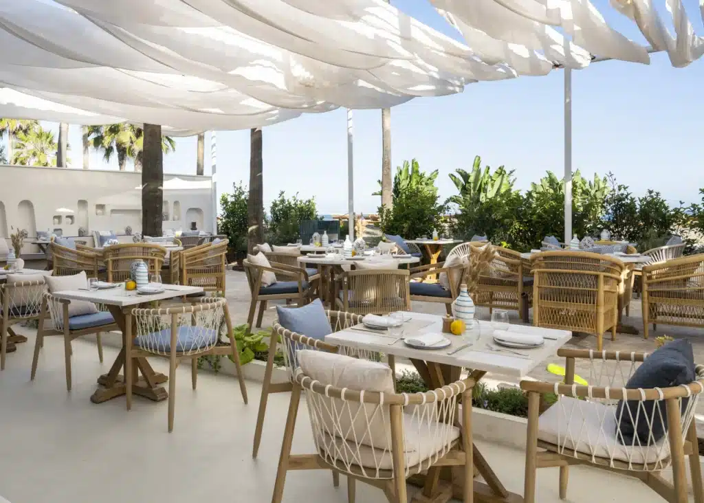 Outdoor restaurant with wicker chairs and wooden tables under white fabric canopies and surrounded by lush greenery.