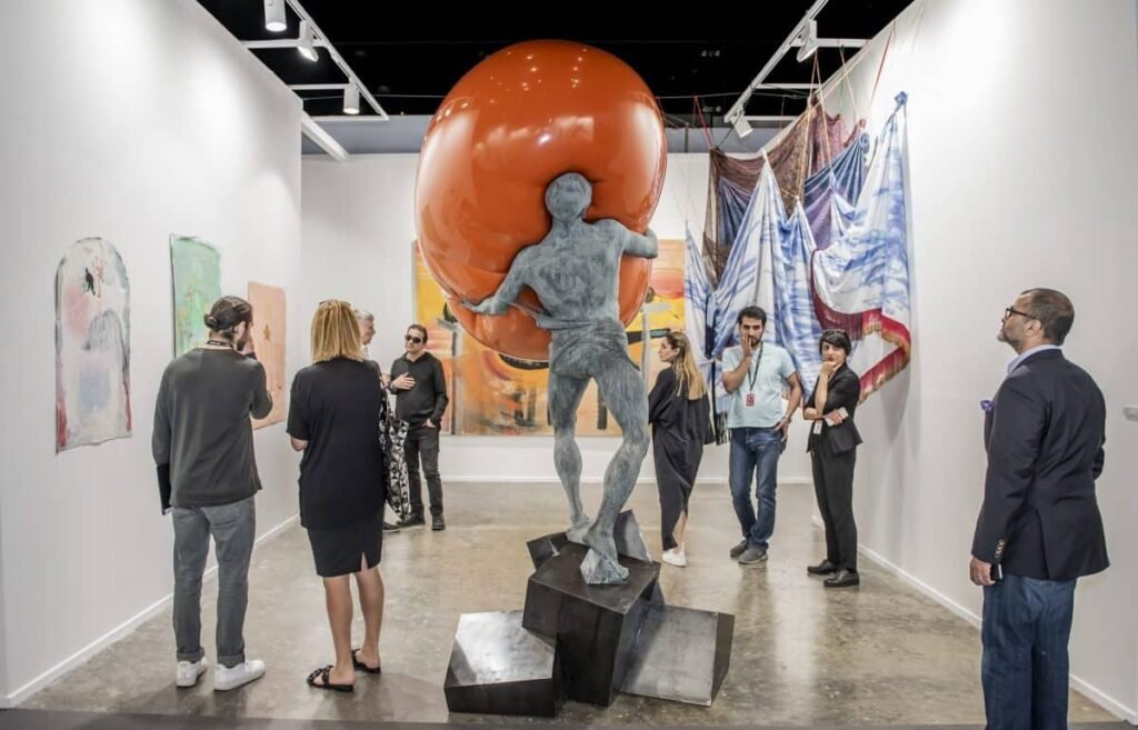 A group of people viewing a contemporary Art Dubai 2026 exhibit featuring a statue of a figure holding an oversized orange sphere.