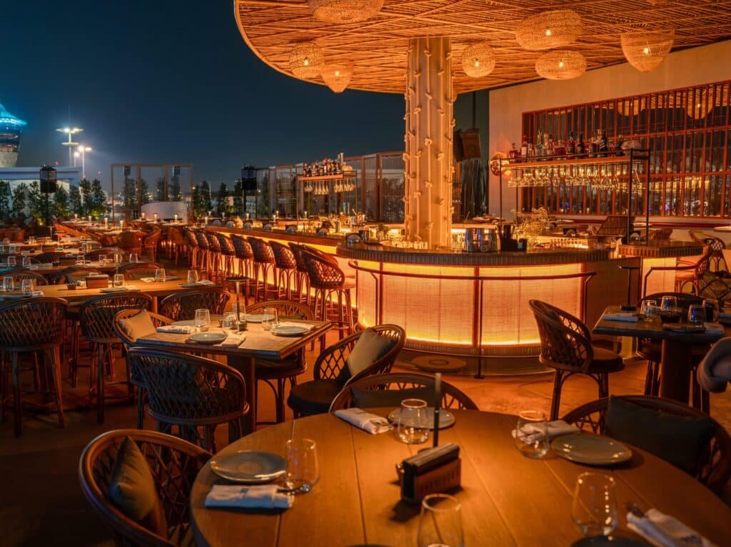 Elegant rooftop restaurant at night, featuring warmly lit tables with neatly arranged place settings and a softly glowing bar.