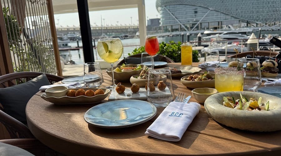 A table set with various dishes and drinks overlooking a marina with boats and a modern building in the background.
