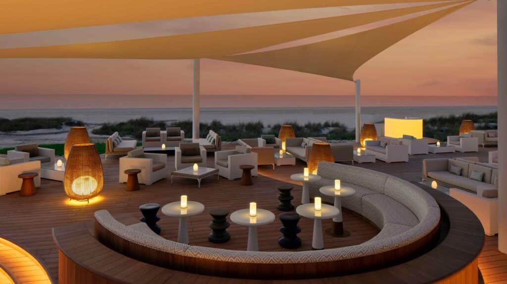 Outdoor lounge with curved seating, tables, and glowing lamps under large sails, overlooking the beach at sunset.