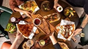 People seated around a table with various dishes, including pizza slices, salads, and sides, sharing a meal together.
