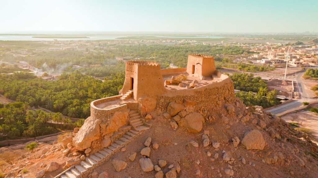 A desert hilltop fort with stone towers overlooks palm groves, a town, and a distant river or lake