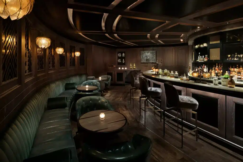Dimly lit bar with leather seating, round tables, a curved wooden counter, and various bottles on display.