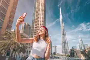 A woman takes a selfie near tall skyscrapers, including the Burj Khalifa, on a sunny day.