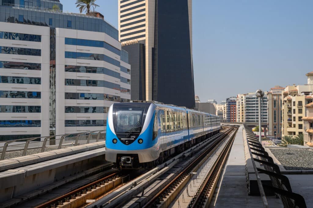 A blue and silver metro train travels on an elevated track between modern high-rise buildings on a clear day.