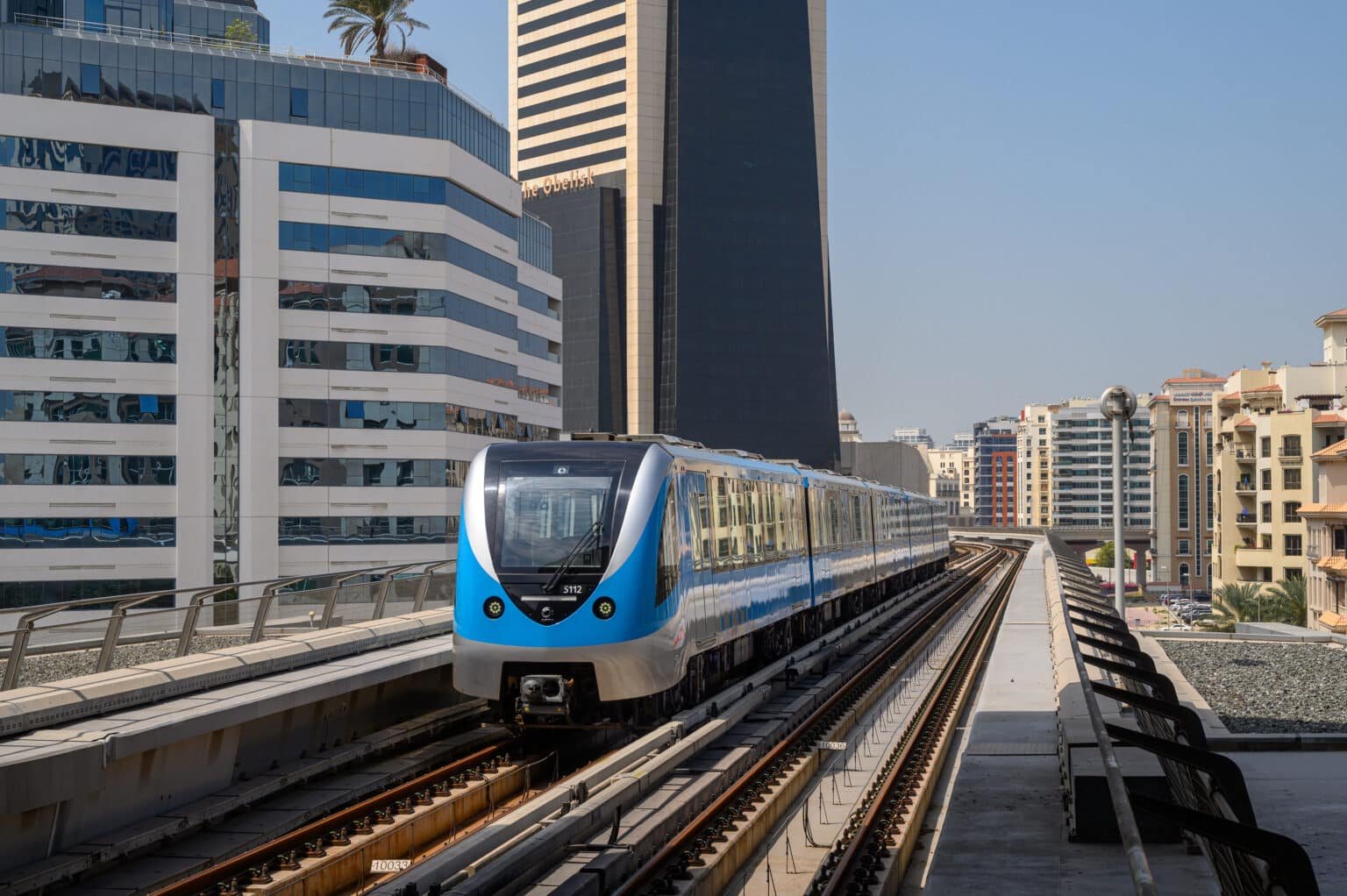 A blue and silver metro train travels on an elevated track between modern high-rise buildings on a clear day.