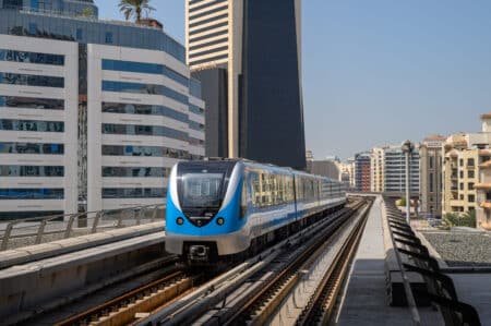 A blue and silver metro train travels on an elevated track between modern high-rise buildings on a clear day.