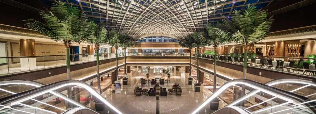 Luxurious hotel atrium with palm trees, escalators, and seating under a patterned glass ceiling
