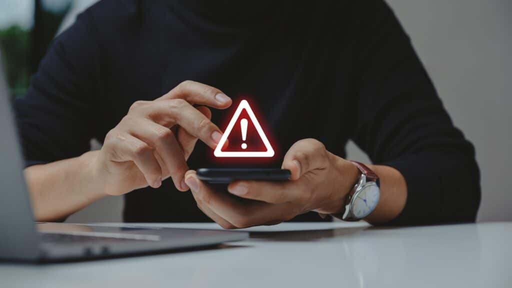 A person holding a smartphone with a glowing warning icon above it, seated at a desk with a laptop.