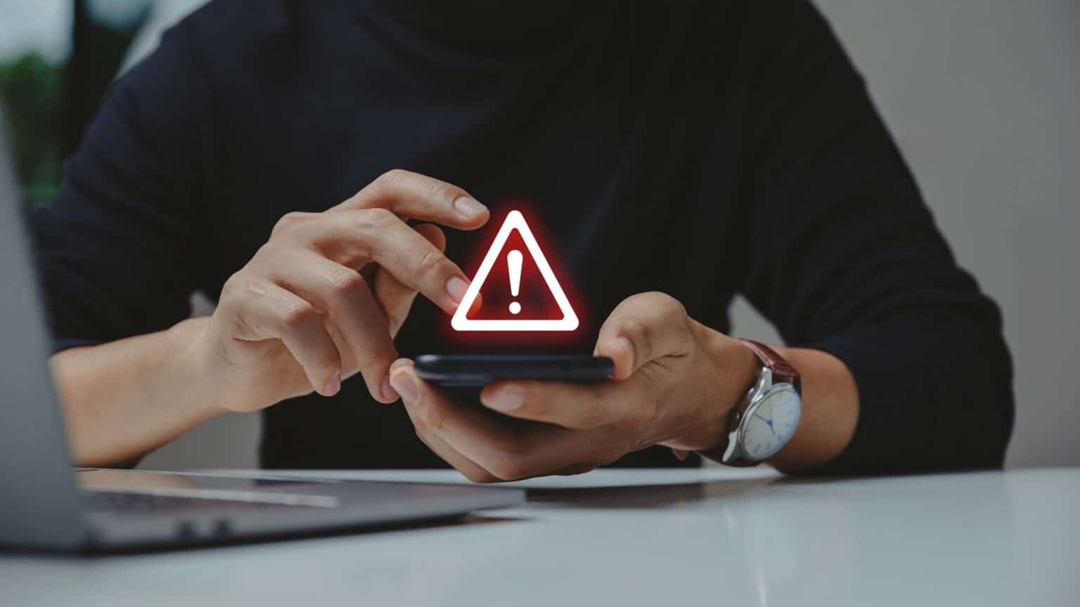 A person holding a smartphone with a glowing warning icon above it, seated at a desk with a laptop.