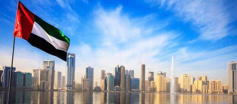 Eid Al Adha UAE flag waving in front of a modern city skyline with skyscrapers and a water fountain.