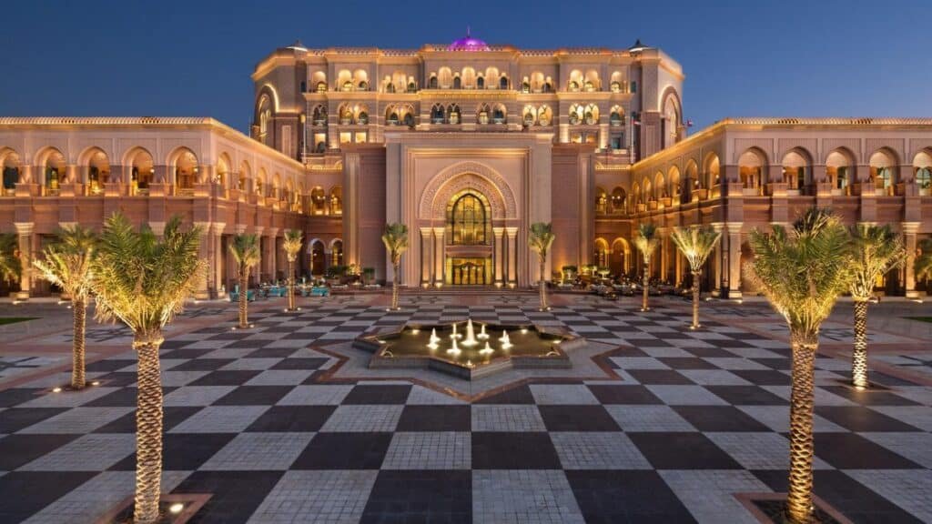 Illuminated grand palace hotel courtyard with palm trees, patterned paving, and a central fountain at dusk