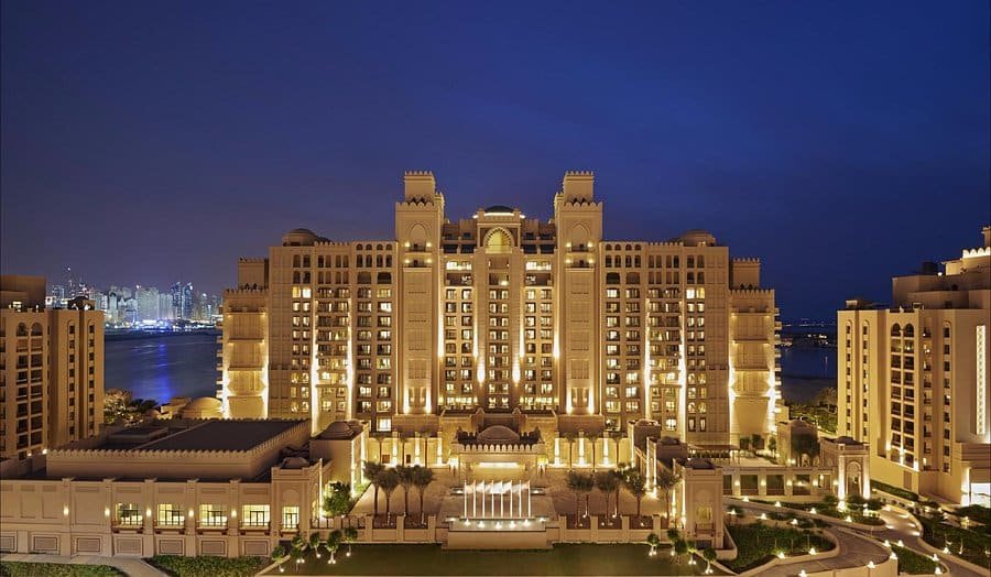 Illuminated luxury hotel exterior with multiple floors and towers, set against a night sky, with a body of water visible in the background.