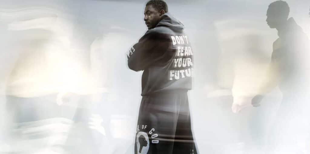 A person wearing a dark jacket with the text Fear of God x Idris Elba