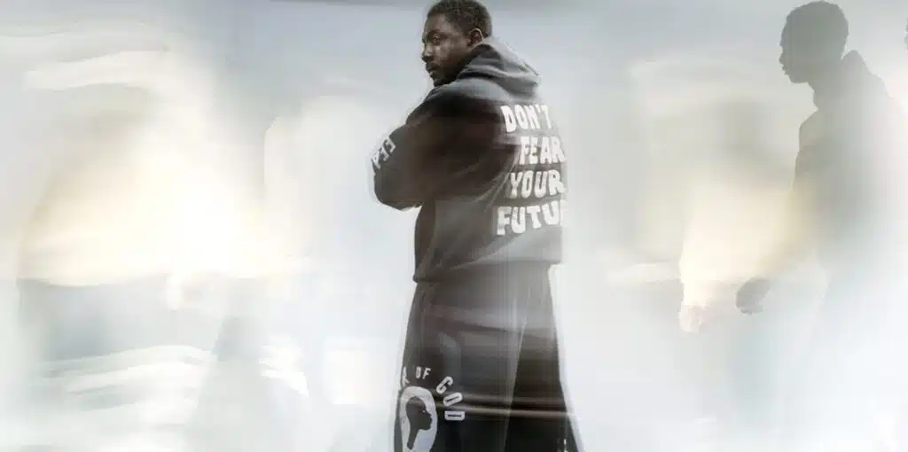 A person wearing a dark jacket with the text Fear of God x Idris Elba