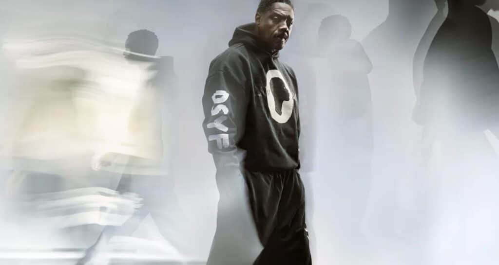 A man in a black hoodie with "OSYF" on the sleeve stands still amid blurred figures in motion.