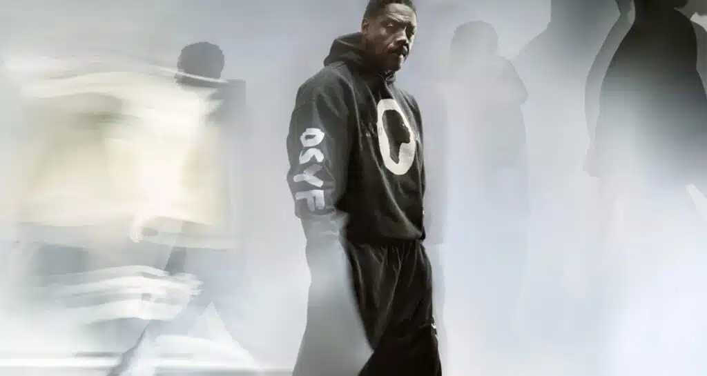 A man in a black hoodie with "OSYF" on the sleeve stands still amid blurred figures in motion.