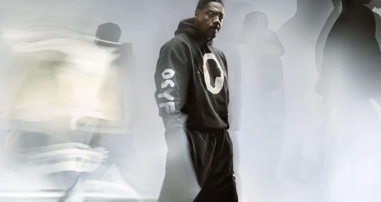 A man in a black hoodie with "OSYF" on the sleeve stands still amid blurred figures in motion.