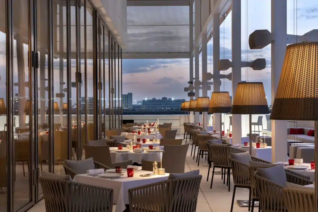 Outdoor rooftop restaurant terrace with set tables, woven chairs, and glowing pendant lamps at dusk