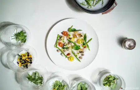 Overhead view of a plated gourmet vegetable dish surrounded by bowls of fresh herbs and edible flowers