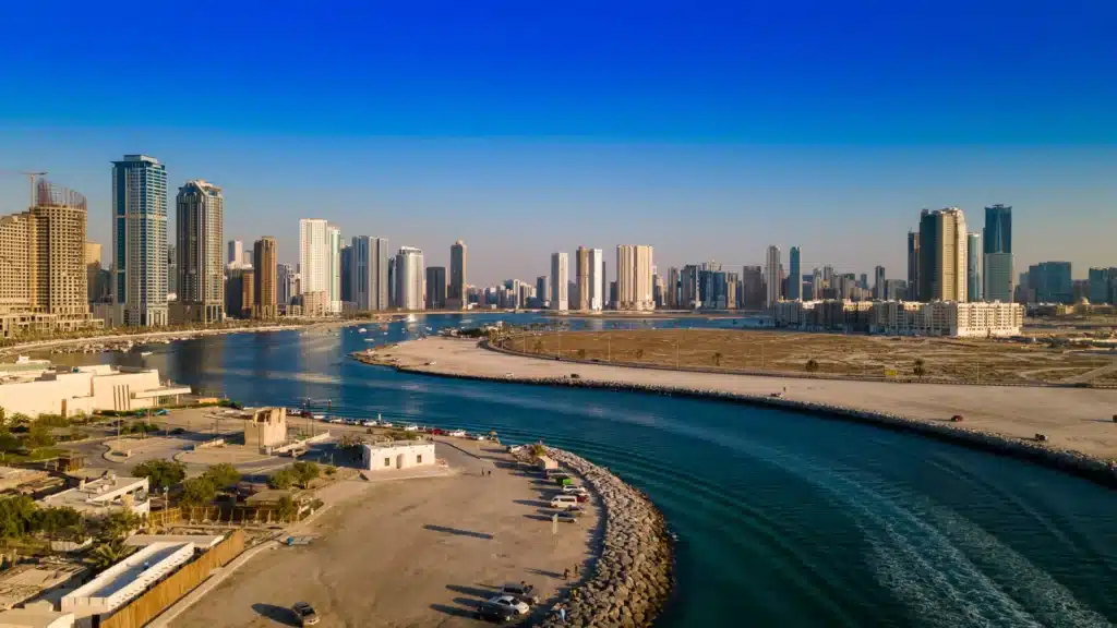 Sunny waterfront city skyline with tall high-rises, a curving channel, and sandy shoreline under a clear blue sky