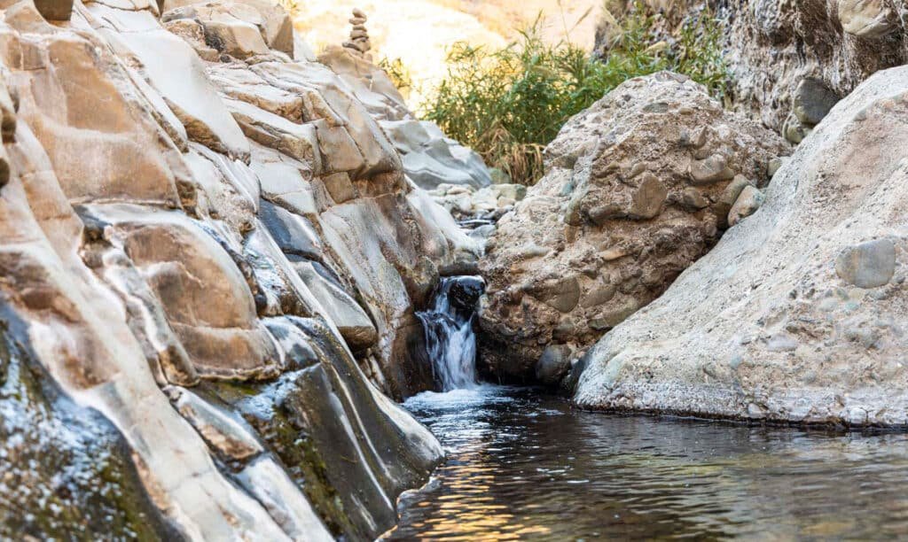 Small waterfall trickling through rocky canyon into a calm pool, with sunlit stone and sparse greenery