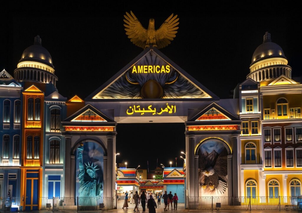 Illuminated entrance gate with “Americas” sign, flanked by vibrant buildings and topped with an eagle statue, at Global Village at night.