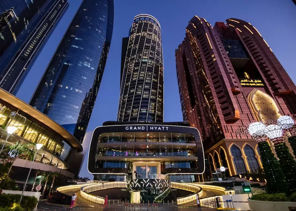 Luxury Staycations In Abu Dhabi: Nighttime view of the Grand Hyatt entrance surrounded by illuminated skyscrapers against a blue sky