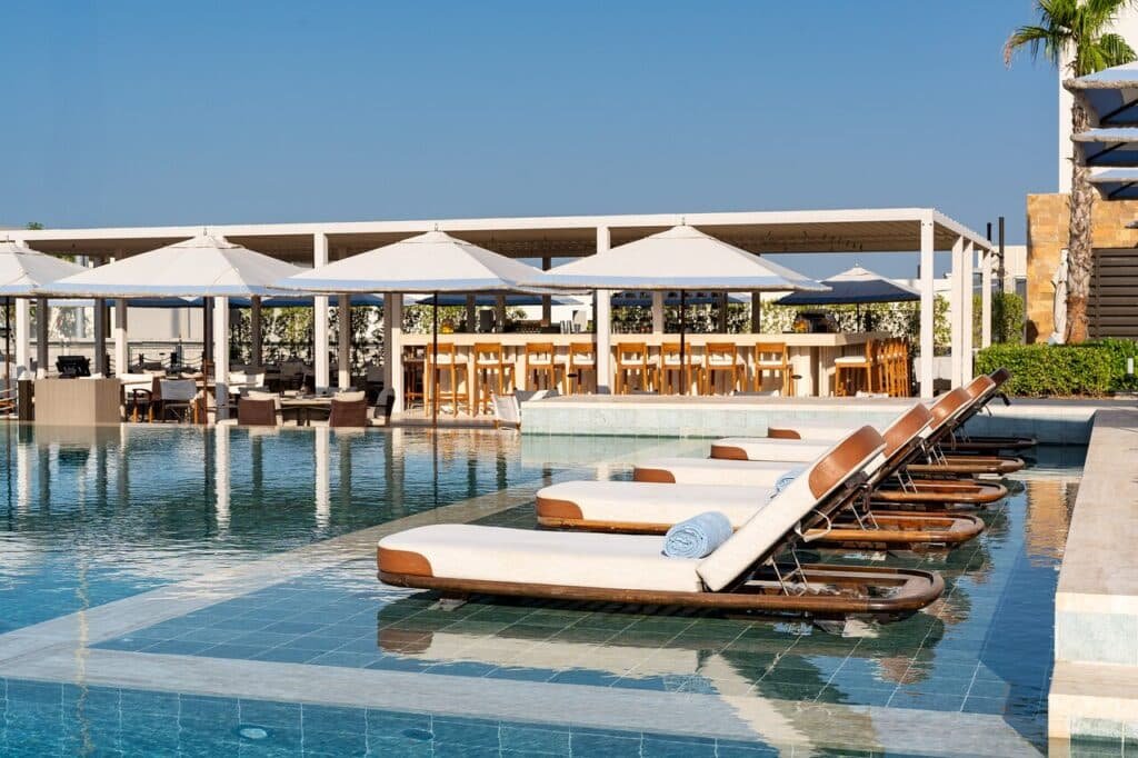 Poolside lounge chairs partially submerged in water, with large white umbrellas and a bar area in the background.