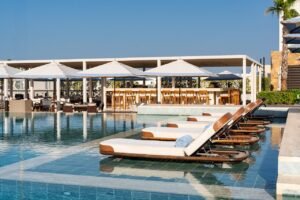 Poolside lounge chairs partially submerged in water, with large white umbrellas and a bar area in the background.