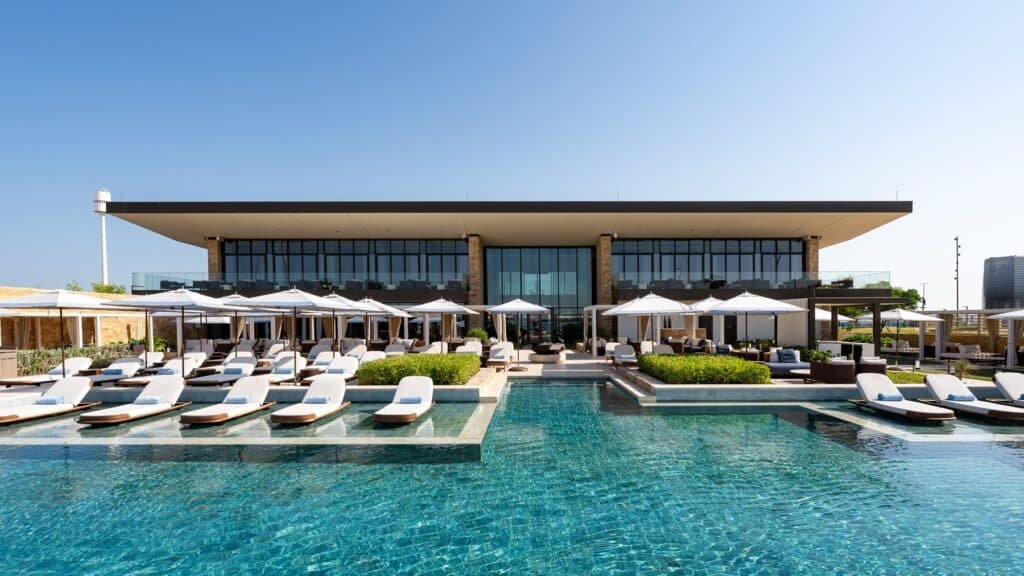 A luxurious Ilios Beach Club swimming pool with white lounge chairs and umbrellas in front of a modern, two-story building under a clear blue sky.